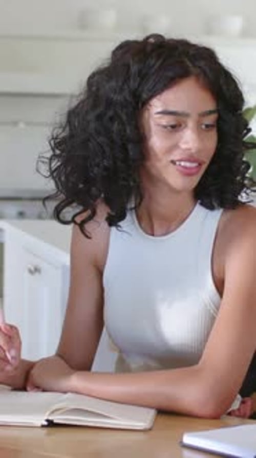 Vertical video: Reacting woman in white in kitchen to offscreen cue, using pink pen near notebook