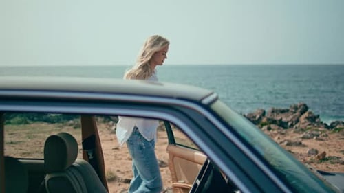 Woman opens car hood after pulling lever on sunny seashore roadside auto trip