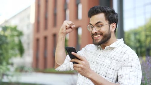 Happy excited man received great news on mobile phone while sitting on a bench on street near modern