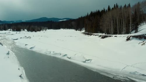 low angle aerial view of river with a white snowy bank and pine trees, Creek.