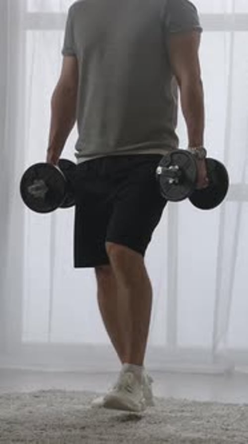 Focused Young Man Performing Home Dumbbell Lunges in a Bright Living Room Demonstrating Lower Body