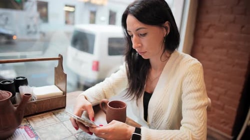 Young Woman Sits in Restaurant or Cafe and Drinks Tea or Coffee From Mug Overlooking City Street
