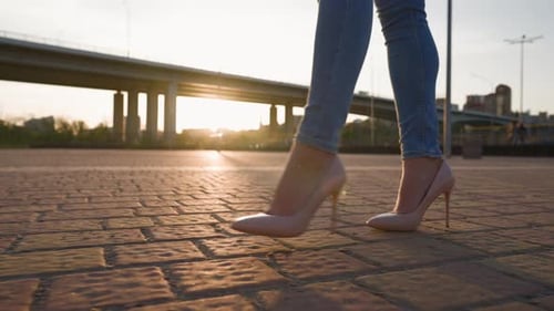 Slim Female Legs in HighHeeled Shoes Walk in the Sunset Light in City Park