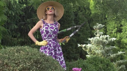 white girl in purple dress and 1950s-style straw hat with sunglasses trims bush with garden shears i