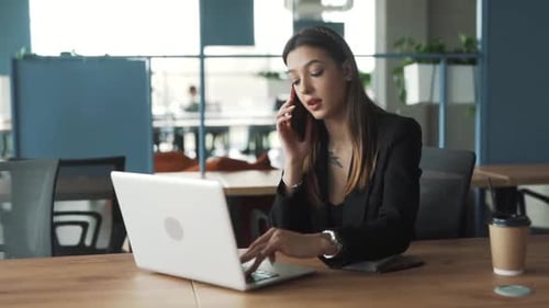A charming woman is seated in the office, working on her laptop and talking on the phone