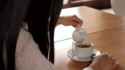 Brunette Girl Gracefully Adds Cream to Her Coffee at a Cozy Cafe