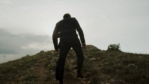 Rear View of an Adult Man in a Business Suit Fluttering in the Wind Climbing Up a Rocky Mountain
