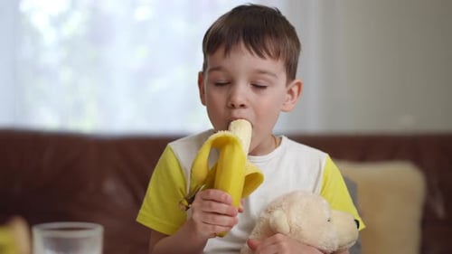 Young Boy Eating Banana at Home with Teddy Bear