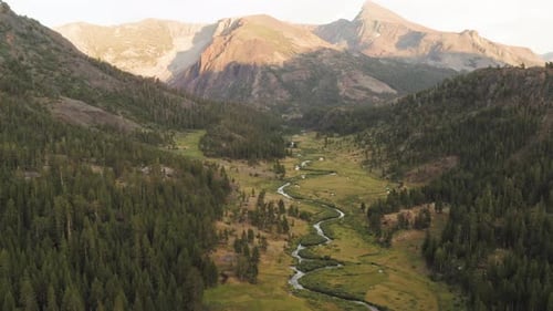 Mountain Valley With Twisting River. Sunlight Hitting Mountain Range In Background. Aerial Flying Ba