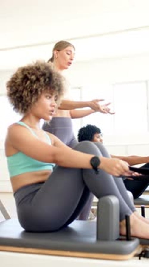 Vertical video: Training on Pilates reformer, fit woman guided by female instructor in yoga studio