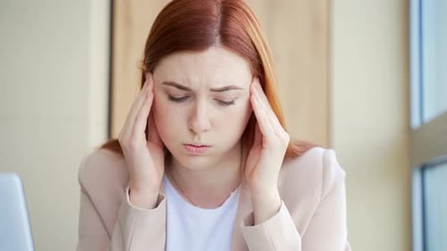 Close-up portrait of a young red-haired woman suffering from a severe headache at work in workplace.