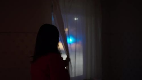 Woman Looks Out the Window of Her House Observing Night Incident Scene with Out of Focus Police