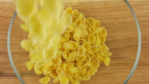 Shell-shaped pasta is poured into a glass transparent bowl against a wooden oak board. Top view.