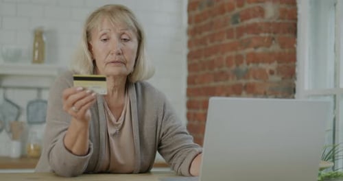 Elderly Woman Paying with Credit Card Using Laptop Shopping Online Indoors in Kitchen in Apartment