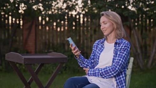 Pleasant Young Woman Sits in the Garden and Uses a Smartphone for Communications Measured Country