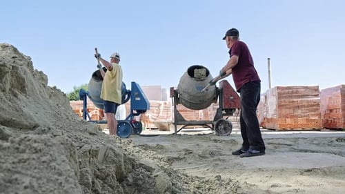 Zwei Bauarbeiter mischen Beton mit Schaufeln und Betonmischer auf umzingelter Baustelle