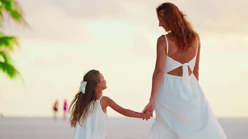 A Beautiful Mother and Her Little Daughter in White Dresses Walk Hand in Hand Along the Ocean Shore