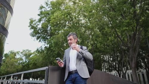 Man Using Phone and Drinking Coffee in City Park
