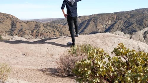 A young man on a hike in the California mountains stands at the top of a cliff overlooking the lands