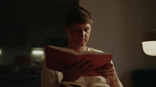 Portrait of Serious Focused Male Reading Paper Book While Sitting on Cozy Sofa in Dimly Lit Room at