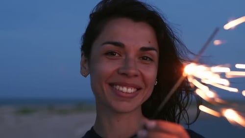 Young Mixed Race Woman Walking Having Fun at Night Party at Seaside with Sparkler in Hand Smiling