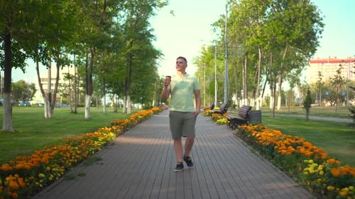 A Young Man Walks in the Park and Drinks Coffee A Man Walks Along an Alley of Flowers with a Glass
