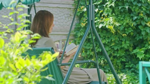 Imágenes comerciales: una niña en verano en el jardín lee un libro sentada en un columpio