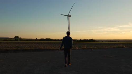 Hombre caminando en el campo cerca de una turbina eólica que genera electricidad, hora dorada