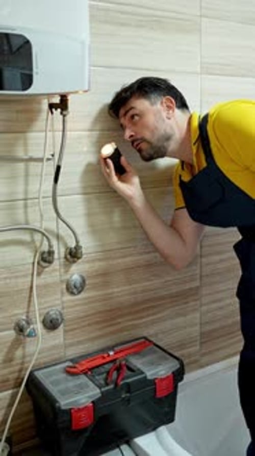 Plumber carefully examining water heater pipes with flashlight, troubleshooting potential issues in