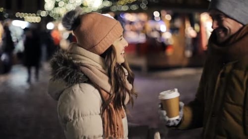 Happy young man brings coffee to his woman at christmas market on winter evening