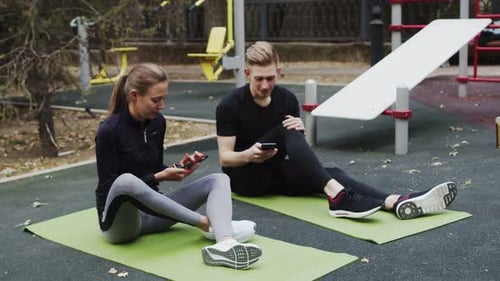 Young Caucasian Sportswoman and Her Trainer Sitting on the Fitness Mats in the Outdoor Workout Area