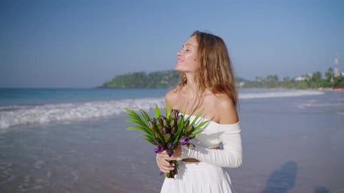 Pretty Woman Smiling and Smelling Bouquet of Purple Flowers at the Ocean Shore Slim Model Walking
