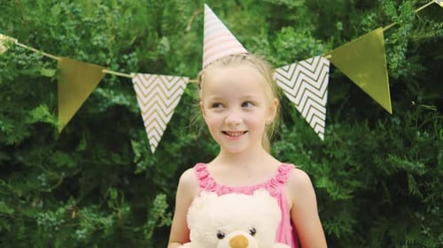 Uma menina em uma camiseta colorida sorrindo com um ursinho de pelúcia em uma festa de aniversário ao ar livre com Happy