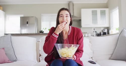 Happy caucasian woman sitting on couch, eating snacks and watching tv in living room