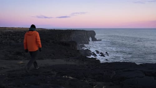 Man In Orange Winter Coat Walking On A Cliff And Looking Towards The Ocean - static shot