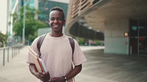Young student stands outside of university, looks at the camera, smiles
