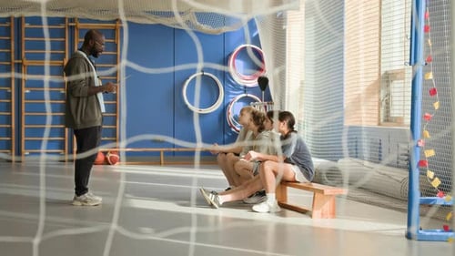 Black Man as Coach Giving Instructions to Children Sitting on Bench in Gym