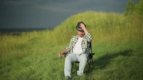 Man Sitting in Field Adjusting Hat with Paintbrush in Hand on a Sunny Day