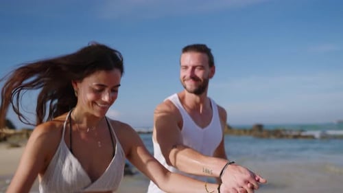 Young Happy Couple Runs Holding Hands on Beach Through Waves Splashing Water and Enjoying Summer
