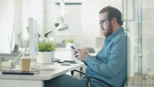 Creative Man Uses Smartphone while Sitting at His Workplace. Office is Light and Modern with Green