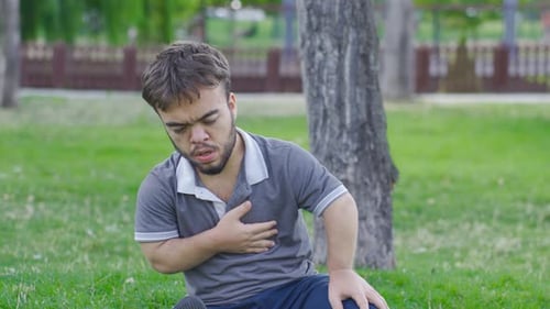 Dwarf young man with chest pain is in the park. Dwarf young man with health problem.