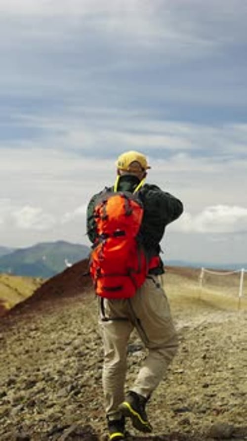 Solo Hiker with Bright Red Backpack Walking Along Rocky Mountain Trail Under Wide Blue Sky Capturing