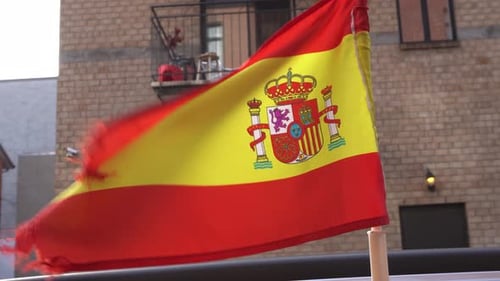 Eine Nahaufnahme der spanischen Flagge und dahinter sieht man ein Gebäude. Die Flagge bewegt sich unter dem Effekt