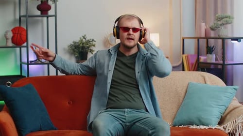 Overjoyed Young Man in Wireless Headphones Dancing Singing on Cozy Couch in Living Room at Home