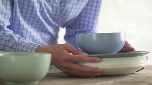A Young Housewife Takes A Stack Of Plates To Set The Table In The Kitchen. A Folded Set Of Clean