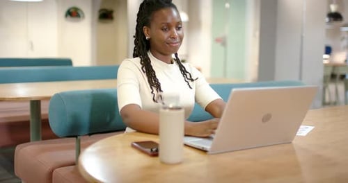 Femme assise à la table commune dans le salon de coworking, commençant à travailler en tapant sur un ordinateur portable avec gobelet