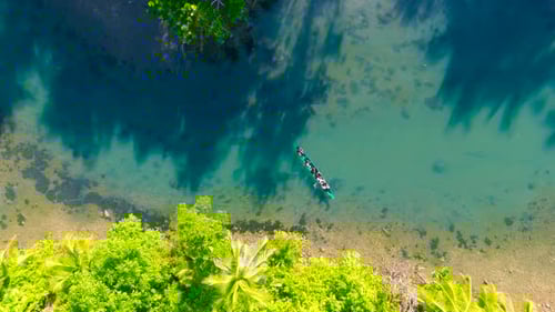 Aerial View of a Long Boat with a Group of People Paddling Through Clear Shallow Water Alongside