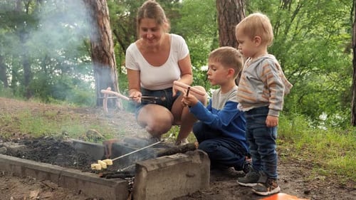 Happy smiling mother with two boys cooking sausages on the bonfire at forest camp
