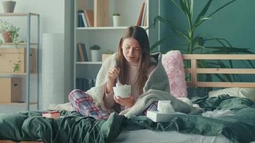 Stressed Young Woman Wrapped in Blanket Sitting on Sofa Eating Ice Cream Crying