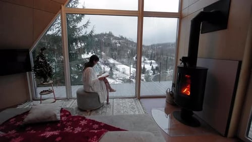 Woman Relaxing By a Fireplace with a Snowy Forest View From a Cozy Room Wrapped in a Robe